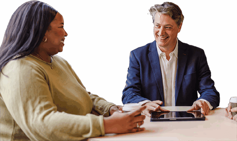 Two people sit at a table, smiling and talking. One person holds a coffee cup, while the other, wearing a suit, has a tablet on the table. Both appear engaged in a friendly conversation.